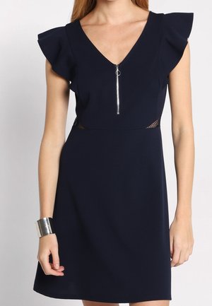 Femme portant une robe bleu marine sans manches avec des détails volantés aux épaules, une fermeture éclair à l'avant et des panneaux latéraux en mesh, accessoirisée d’un bracelet manchette argenté.
