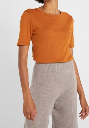 Mujer con una blusa ajustada de manga corta color naranja quemado y pantalones de punto gris claro de cintura alta con una banda elástica acanalada.