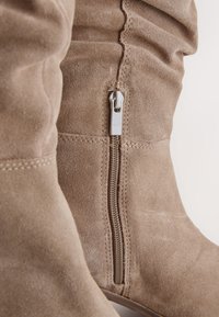 Bottes hautes en daim couleur fauve avec un détail décoratif de fermeture éclair sur le côté. Texture douce et accents cousus qui renforcent le design général.