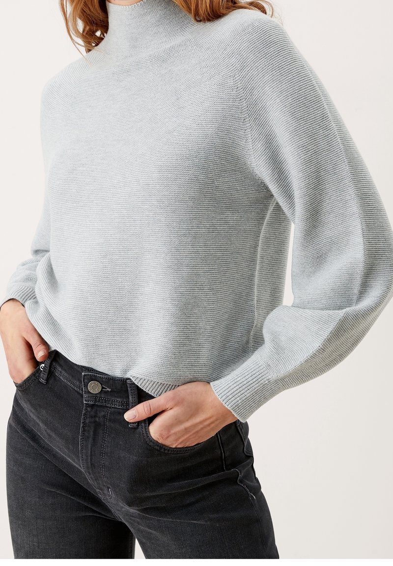 Femme portant un pull à col roulé côtelé gris clair avec des manches bouffantes et un jean noir taille haute, les mains dans les poches.