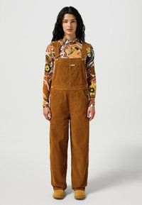 Brown corduroy overalls with adjustable straps, front pockets, and a relaxed fit. Paired with a colorful, patterned long-sleeve shirt.