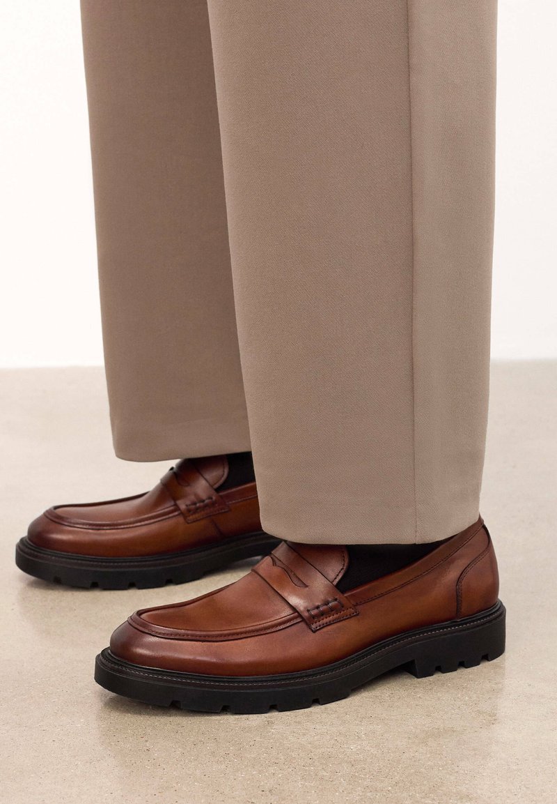 Mocasines de cuero marrón con suelas de goma negras, con detalles de costura y un acabado suave, llevados con pantalones beiges.