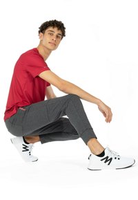 Rød kortærmet træningsshirt, grå teksturerede joggerbukser og hvide sneakers med sorte detaljer. Modellen er i en crouched position.