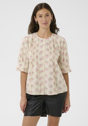 Femme portant un chemisier crème à motifs floraux avec des manches bouffantes et un short en cuir noir, debout devant un fond blanc uni.