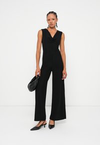 Mono negro con cuello de urna, diseño sin mangas y pantalones de pierna ancha, combinado con un bolso negro y tacones de punta.