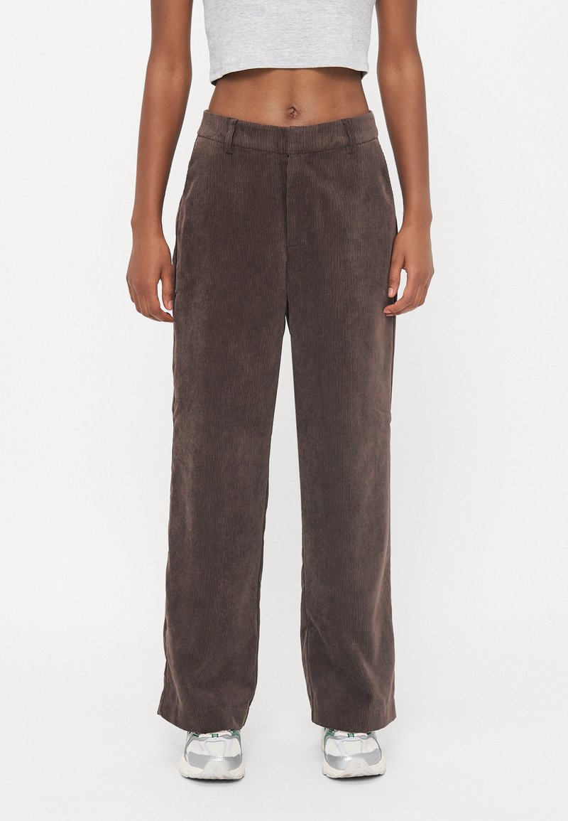 I pantaloni a gamba larga in corduroy marrone presentano una texture a costine, una vita media e sono abbinati a sneakers grigie.