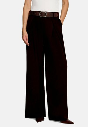 Femme portant un pantalon noir à jambes larges avec des plis devant, une ceinture brun foncé texturée, un haut blanc et des chaussures à bout pointu, debout avec une main dans la poche.