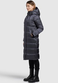 Abrigo largo acolchado en gris oscuro con capucha, que presenta un diseño acolchado y bolsillos laterales. Combinado con pantalones negros y botas de tobillo.