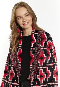 Gehaakte poncho in zwart, rood en wit geometrisch patroon, met franjes aan de randen en een open voorkant. Draag het over een zwart shirt.
