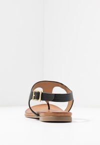 Sandalia de cuero negro con una correa texturizada, detalle de hebilla dorada y suela plana color caramelo; diseño de parte trasera abierta; estilo minimalista.