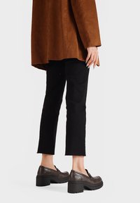 Giacca oversize in suede marrone, pantaloni neri a vita alta, e mocassini con tacco marrone scuro e suola spessa con dettagli decorativi.