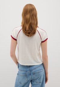 Witte top met korte mouwen en rode accenten, met een gestructureerd ontwerp en een afgeronde onderrand, gecombineerd met hoog-getailleerde blauwe spijkerbroeken.