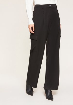Personne portant un pantalon cargo noir à jambes larges avec poches latérales à rabat et des bottines noires à talons pointus, associé à un haut blanc à manches longues.