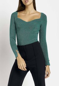 Grön ribbad långärmad bodysuit med fyrkantig halsringning och dekorativa knappdetaljer vid ärmarnas ändar, i kombination med svarta högmidjade byxor.