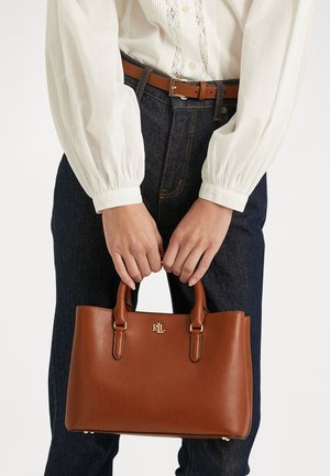 Ręce trzymające brązową skórzaną torebkę z krótkimi uchwytami, złotymi elementami i logo RLL, noszoną z białą bluzką i ciemnoniebieskimi dżinsami.