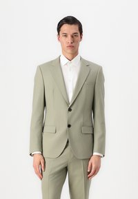 Traje verde claro a medida con chaqueta de un solo botón, botones negros, solapas con muesca y pantalones ajustados a juego. Camisa blanca por debajo.