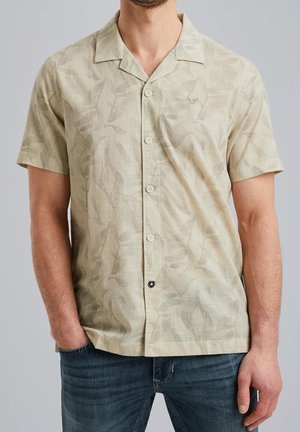Homme portant une chemise beige à manches courtes boutonnée avec un motif discret de feuilles et un col, associée à un jean bleu sur un fond uni.