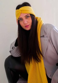 Un bandeau et une écharpe en tricot jaunes, portés avec un manteau en cuir synthétique gris clair. L'écharpe a une texture douce, tandis que le bandeau présente un design torsadé.