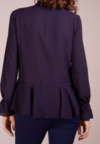 Marinblus med röda prickar, långärmad och med peplumslut. Tillverkad av lättviktsmaterial, med en subtil lyster och ihopdragna manschetter.