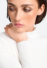 Weißer, gerippter Rollkragenpullover mit texturiertem Finish. Nahaufnahme des Gesichts eines Models, das ein natürliches Make-up und kurze Haare zeigt.