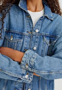 Osoba s kudrnatými vlasy má zkřížené ruce a nosí světle modrou džínovou bundu přes bílý žebrovaný top, přičemž ukazuje detaily bundy a knoflíky.
