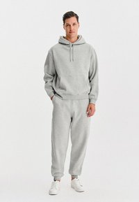 Mężczyzna w jasnoszarej bluzie z kapturem i pasujących dresach, w białych sneakersach, na jednolitym białym tle.