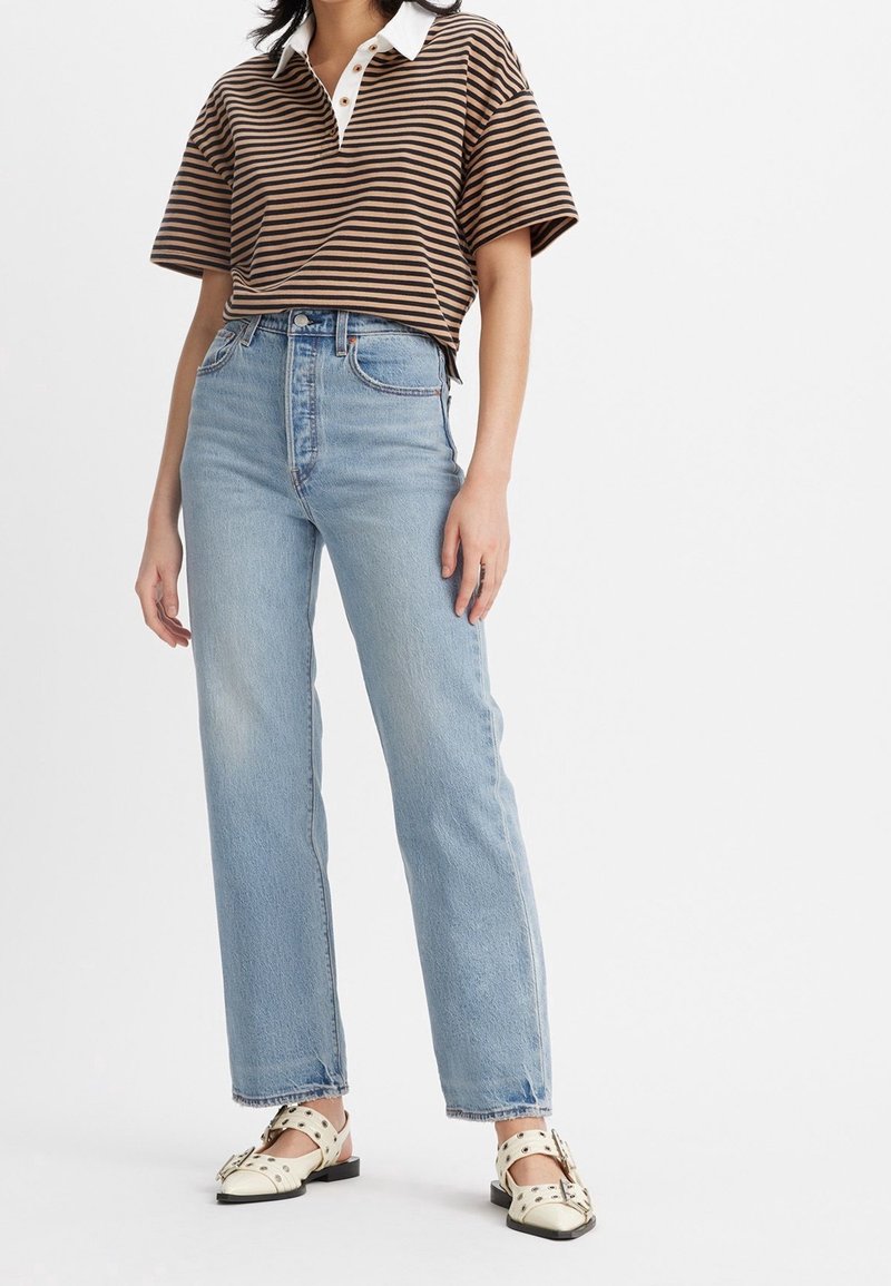 Personne portant un jean taille haute bleu clair, un polo à manches courtes rayé marron et noir, et des chaussures plates blanches cloutées.