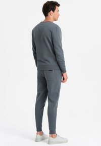 Grauer Kapuzenpullover und passende Jogginghose aus einem weichen, strukturierten Stoff. Die Jogginghose hat eine Gesäßtasche und gerippte Bündchen und wird mit hellen Schuhen kombiniert.
