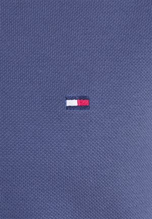 Close-up af blå strikstof med et lille rektangulært logo broderet i hvide, røde og marineblå tråde i midten.