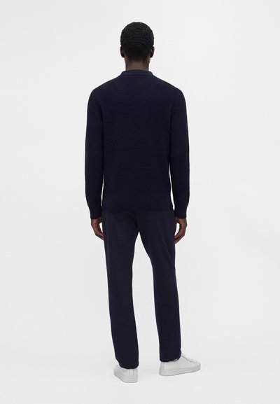 Homme debout, de dos, portant une chemise à manches longues sombre et texturée, un pantalon foncé et des baskets blanches sur un fond uni.