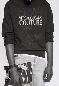 Černá mikina s nápisem "VERSACE JEANS COUTURE" v kontrastní bílé barvě a tónovém vyšívání. Model drží vzorovanou kabelku s designem řetězu.