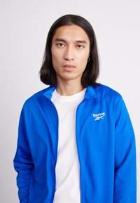 Casaco azul com fecho éclair da Reebok, com um logótipo branco, tecido leve, gola alta e textura suave, usado sobre uma camisa branca.