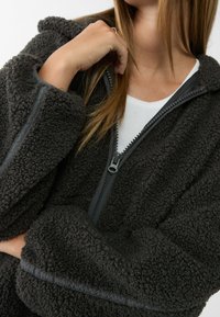 Vezelachtige donkergrijze ritjas met een hoge kraag, voorzien van een gladde ritssluiting en elastische accenten op de mouwen en de zoom.