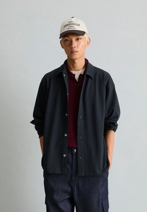 Junger Mann, der eine weiße Kappe, ein kastanienbraunes Hemd, ein schwarzes Hemd mit Knöpfen und eine dunkelblaue Hose trägt, steht mit den Händen in den Taschen vor einem grauen Hintergrund.