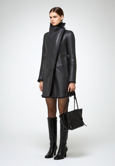 Schwarzer Ledermantel mit hohem Kragen und Kunstfellbesatz, der eine schräg geschnittene Design hat, mit Reißverschlusstaschen, kombiniert mit knielangen Lederstiefeln und einer Handtasche.