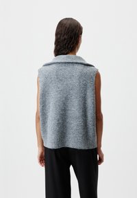 Person mit nassen, dunklen Haaren, der einen ärmellosen, hellblauen Strickpullover und schwarze Hosen trägt, von hinten vor einem weißen Hintergrund gezeigt.