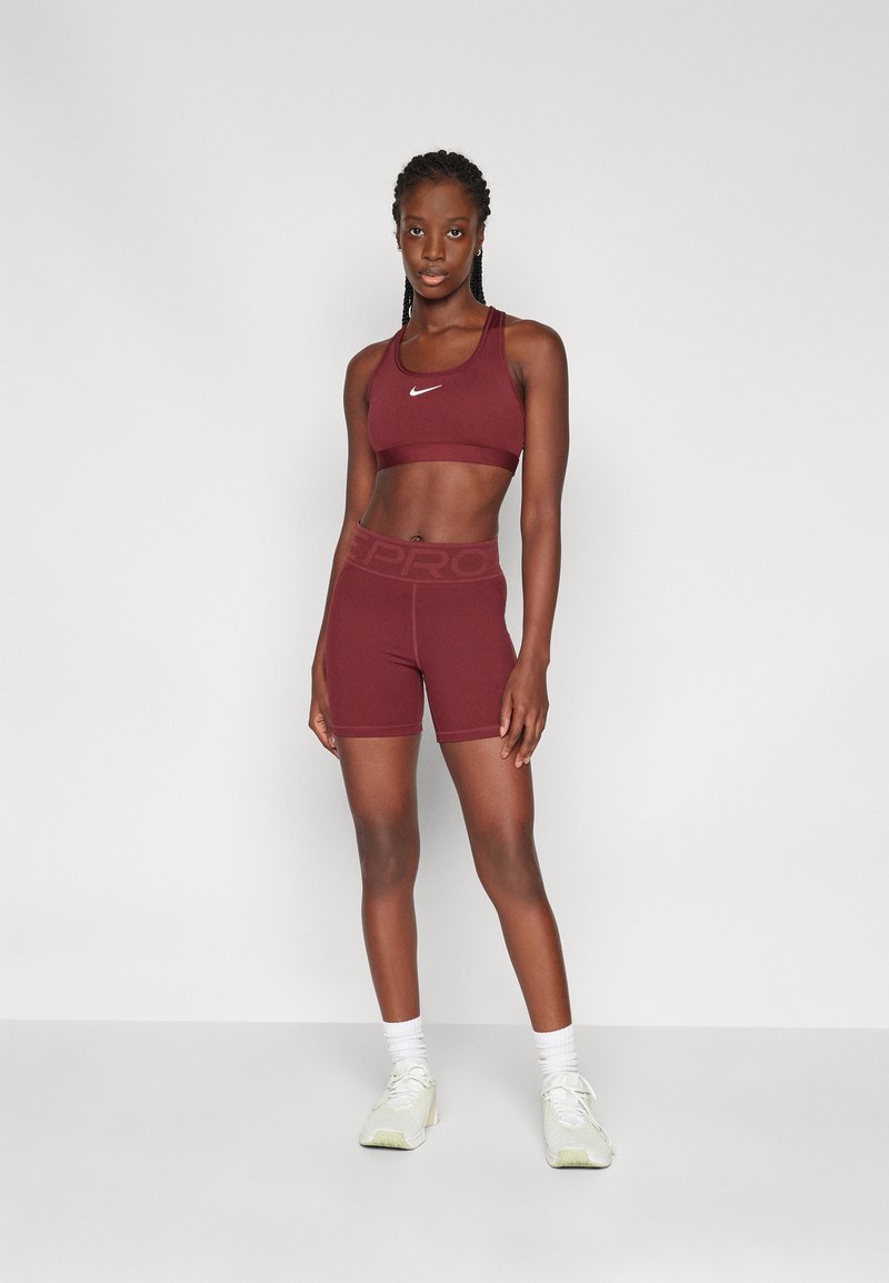 Reggiseno sportivo color mogano e pantaloni corti, realizzati in materiale elastico. Il reggiseno presenta spalline regolabili e un logo bianco. I pantaloni corti hanno una vita larga con branding.