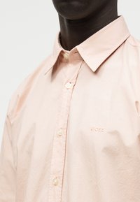 Άτομο που φορά ένα ανοιχτό ροζ πουκάμισο με κουμπιά και γιακά, με την λέξη "BOSS" κεντημένη στο αριστερό στήθος, σε μονόχρωμο φόντο.