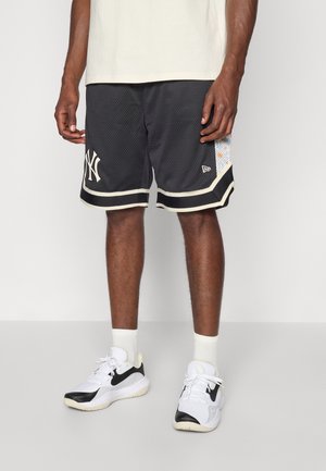Homme portant un short de basket noir des New York Yankees, des baskets blanches Nike et des chaussettes blanches, debout sur un sol gris avec un fond blanc.