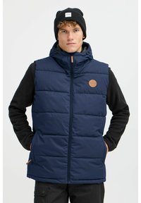 Marineblauer Puffer-Weste mit einem Reißverschluss, gestepptem Design und einem sandfarbenen Logo-Patch. Getragen über einem schwarzen Langarmoberteil.