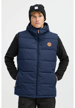 Marineblauer Puffer-Weste mit einem Reißverschluss, gestepptem Design und einem sandfarbenen Logo-Patch. Getragen über einem schwarzen Langarmoberteil.