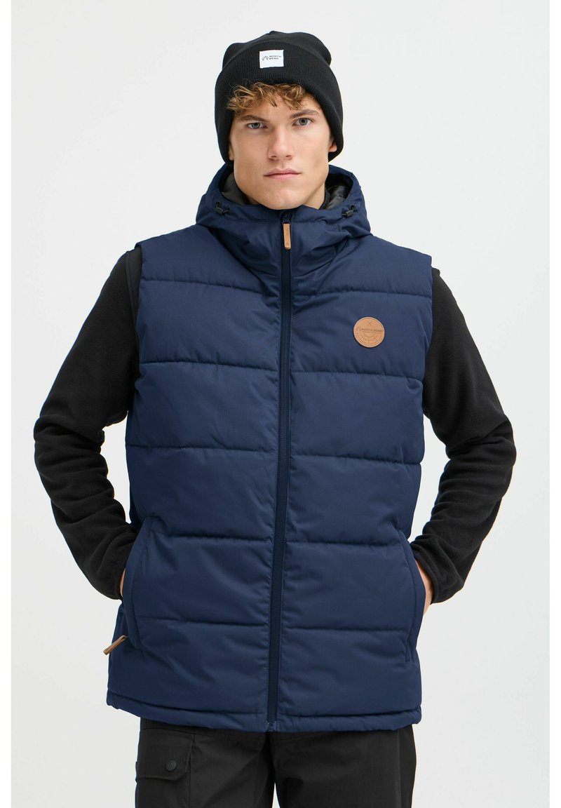 Marineblauer Puffer-Weste mit einem Reißverschluss, gestepptem Design und einem sandfarbenen Logo-Patch. Getragen über einem schwarzen Langarmoberteil.