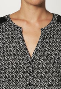Blouse à motifs noir et blanc avec un design en diamant, dotée d'un décolleté en V, de fermetures à boutons et d'un tissu texturé avec des accents noirs sur les épaules.