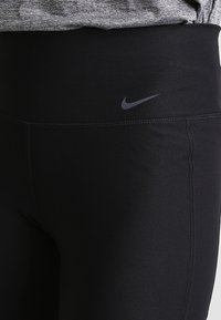 Zbliżenie czarnych legginsów Nike z wysokim stanem noszonych z szarym topem, pokazujące szwy i małe logo Nike w kształcie łuku.