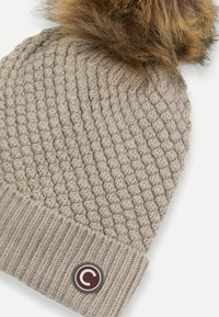 Berretto lavorato a maglia di un beige chiaro con bordo a coste, caratterizzato da un soffice pom-pom in pelliccia sintetica sulla parte superiore e una toppa rotonda con logo sul lato.
