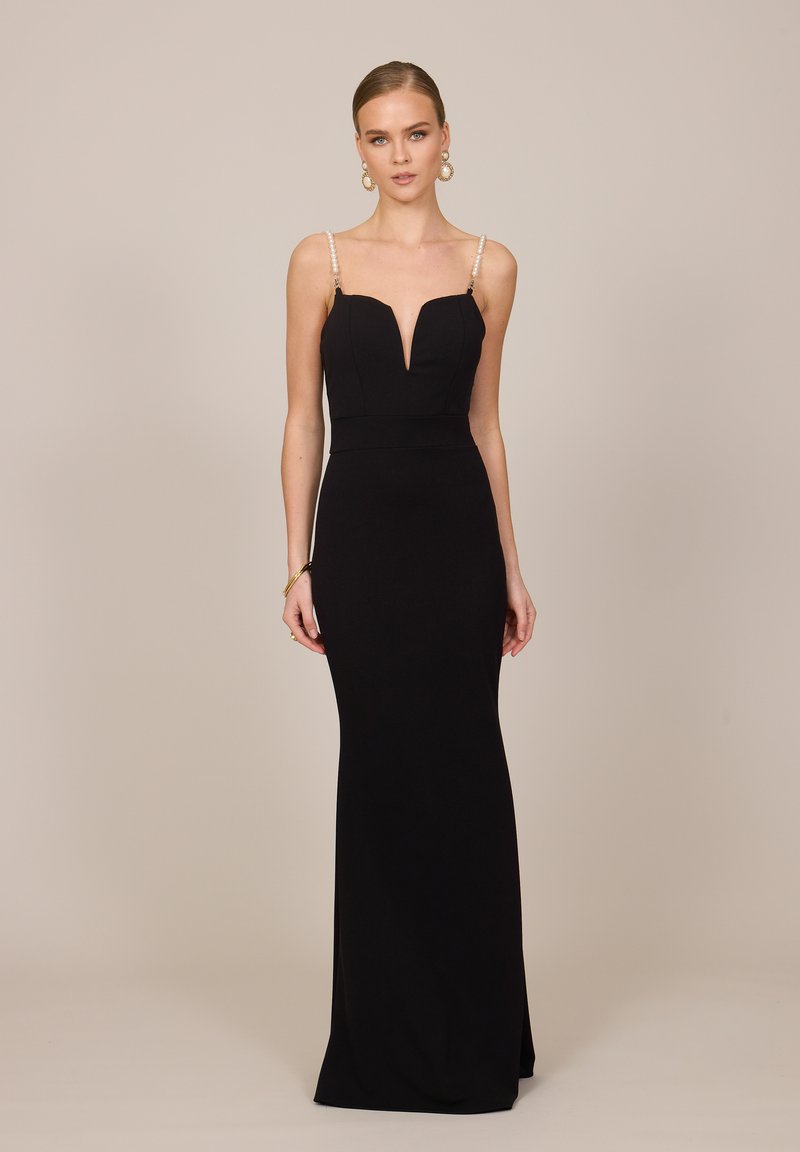 Femme portant une longue robe noire sans manches avec des bretelles en perles et un décolleté en V, debout sur un fond beige uni.