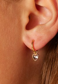 Boucle d'oreille en or avec un pendentif en forme de cœur transparent. Texture lisse, finition brillante et un petit détail de fermoir visible.
