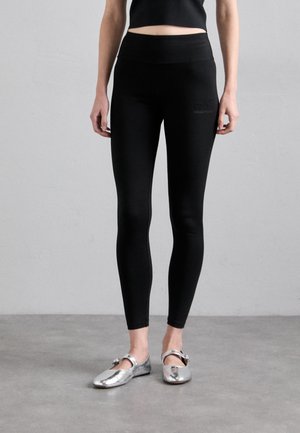 Zwarte leggings met een hoge taille, een gladde textuur en een subtiele logodetail op de bovenbeen. Gekoppeld met glanzende zilveren ballerina's.