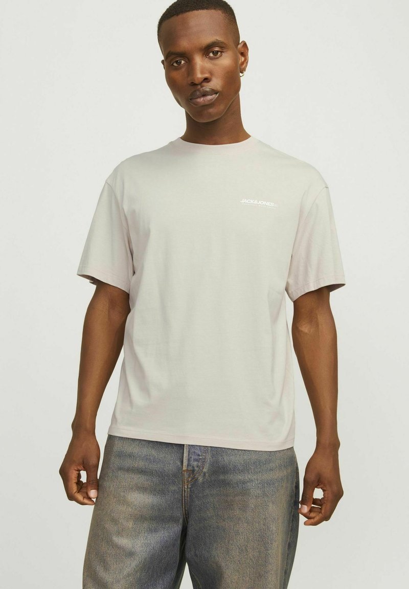 Lys grå bomulds t-shirt med korte ærmer, rund hals og et lille hvidt logo på brystet. Bæret med blå denim jeans.