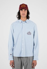 Gestreiftes Langarmhemd in Blau und Weiß mit einer Brusttasche und rotem Sticklogo. Burgunderfarbene Kappe mit goldener Schrift und Akzenten.